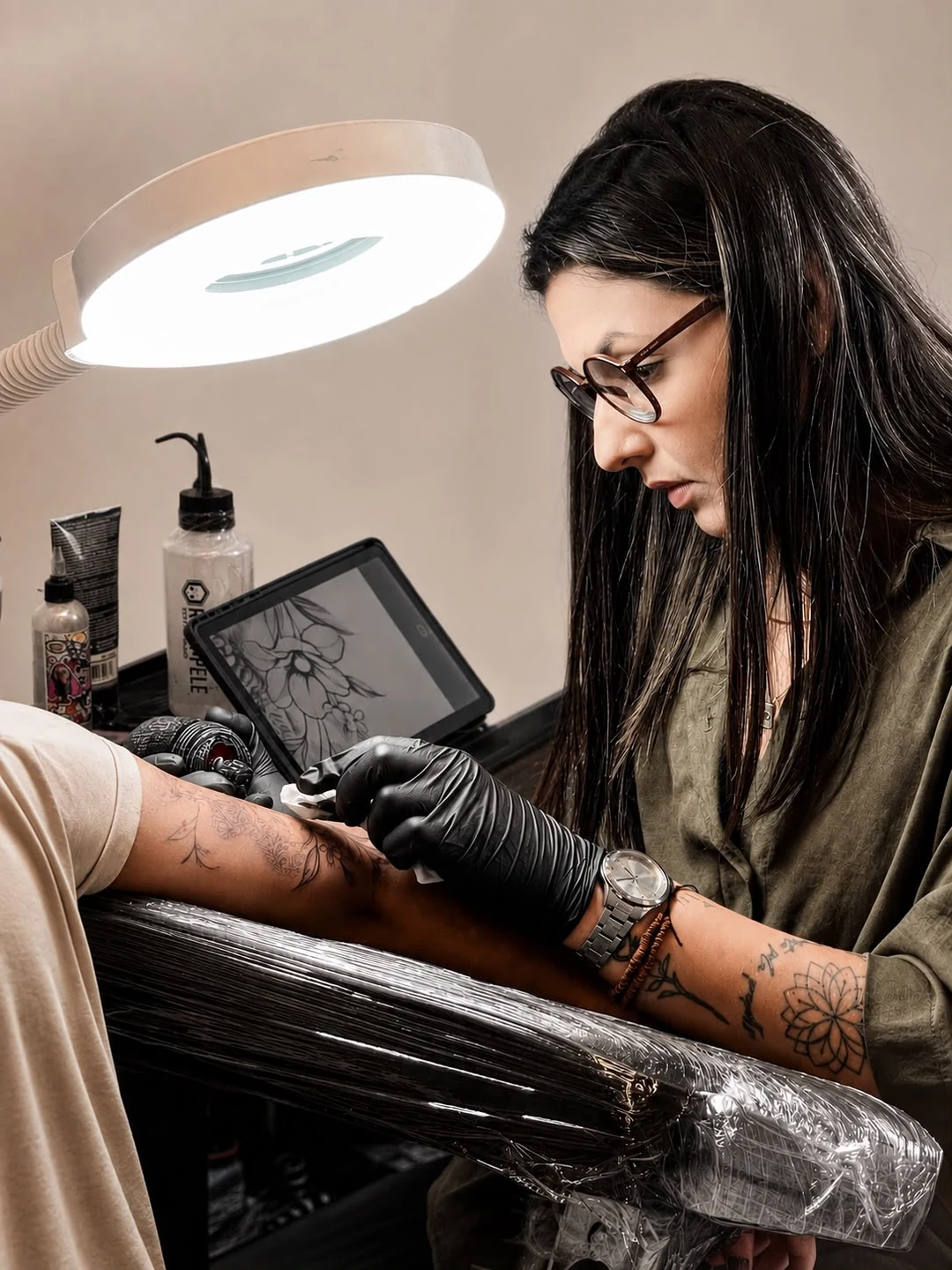 Thay tatuando uma cliente, com projeto no tablet e luminária profissional no estúdio